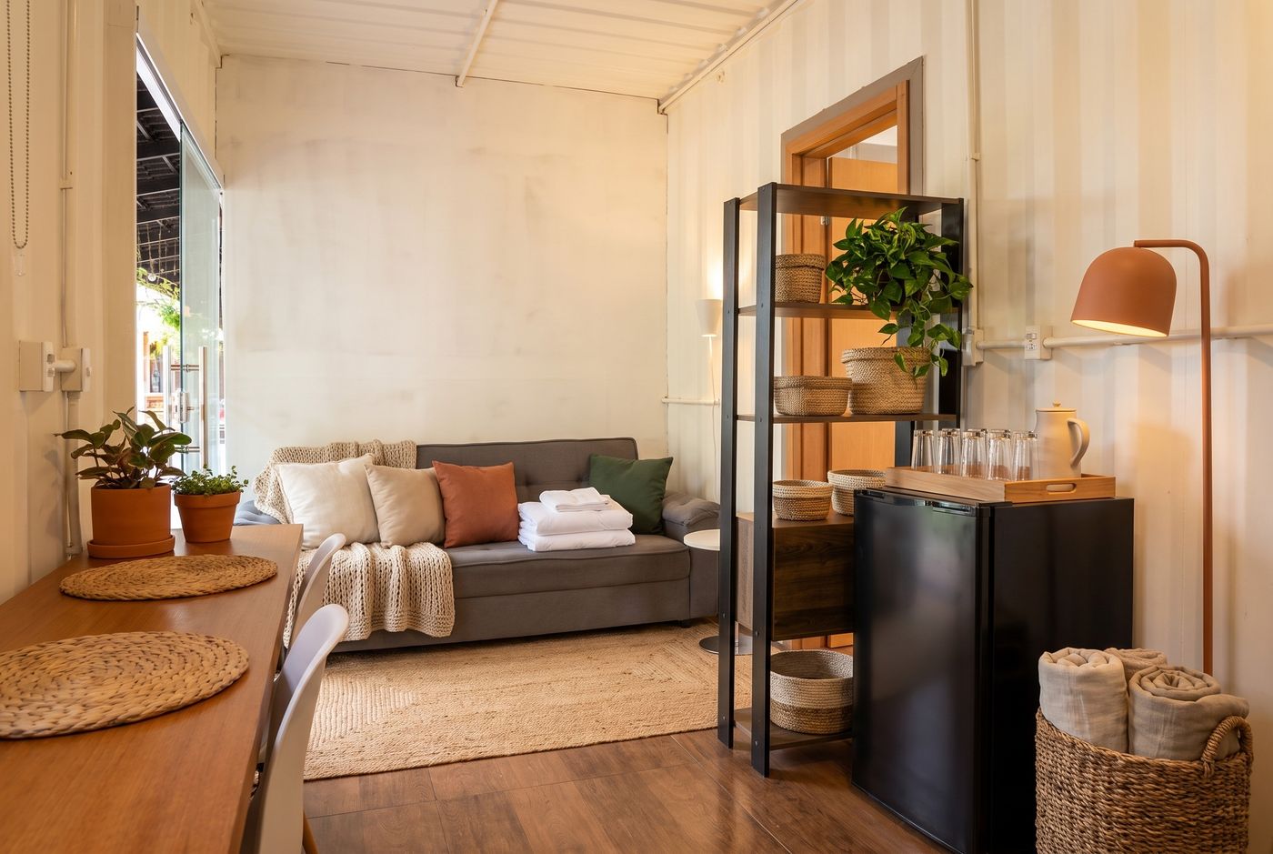 A sala-quarto pelo lado do sofá-cama: estofado de cor média, almofadas lisas, fibra nos cestos e no tapete, e o frigobar ao lado da estante. Imagem conceitual gerada por IA.